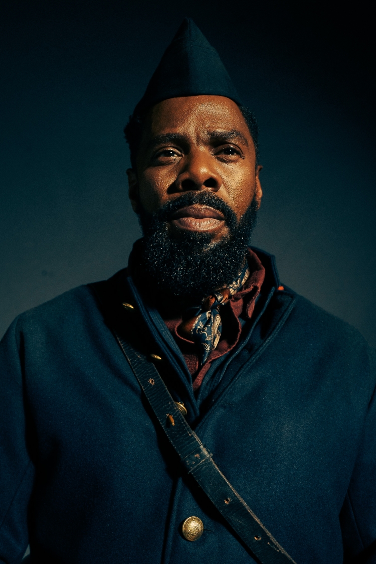 Portrait of a Black man with a beard in a military uniform with a neckerchief and dark hat on a blue background.