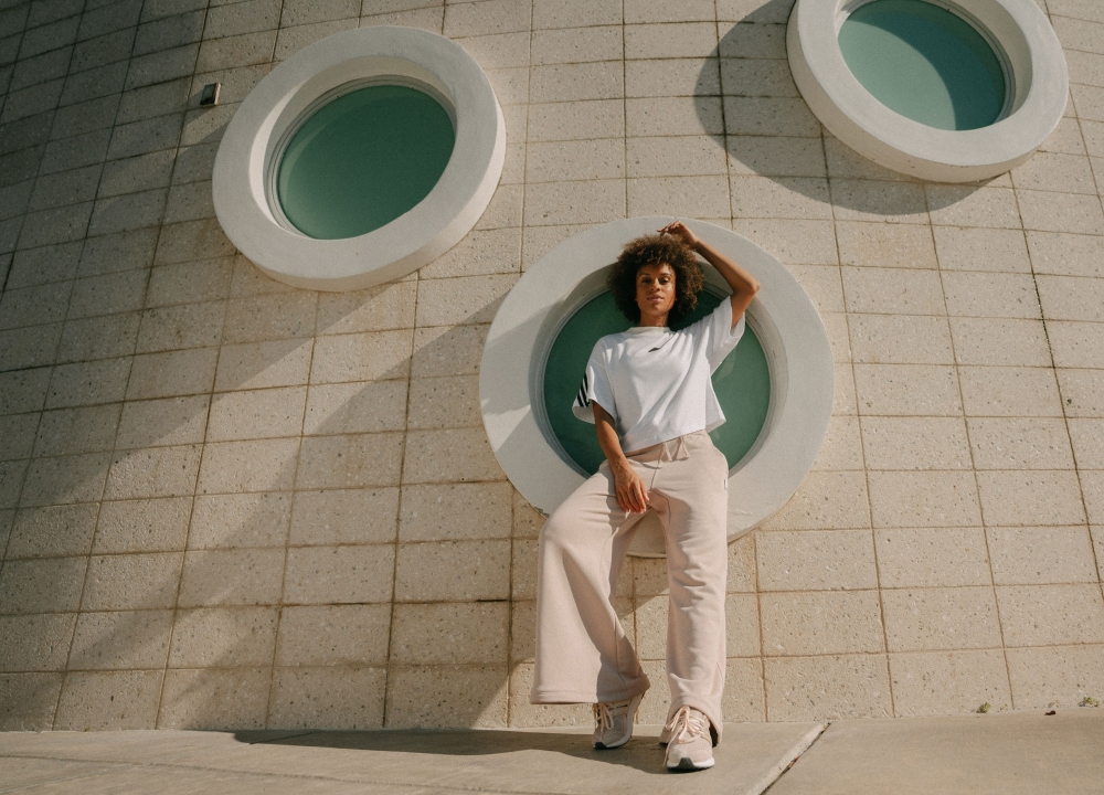 A curly-haired woman poses in wide-leg trousers and an Adidas t-shirt in front of a round window in a modern building on a sunny day.
