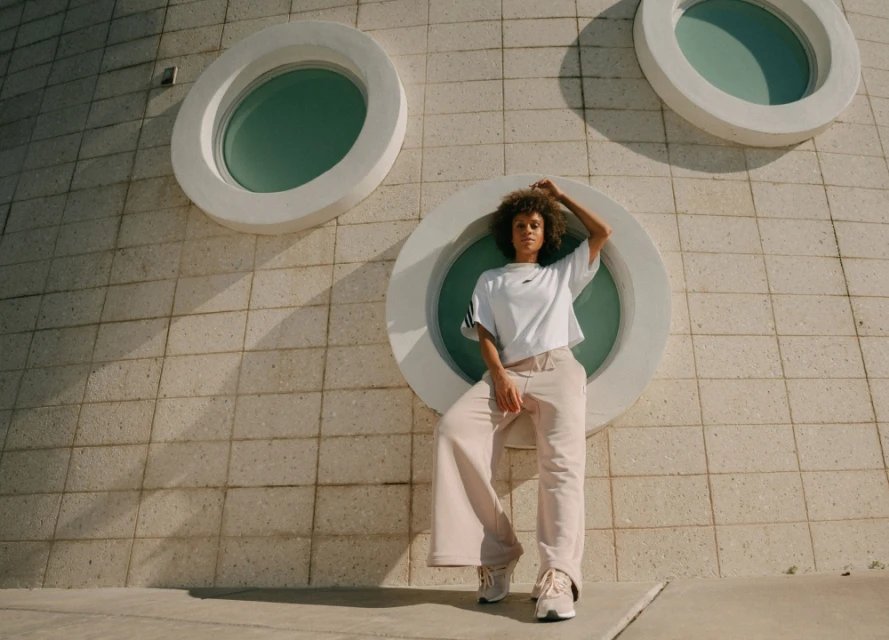 A curly-haired woman poses in wide-leg trousers and an Adidas t-shirt in front of a round window in a modern building on a sunny day.