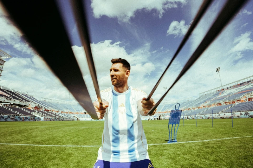 Athlete Lionel Messi training with resistance bands on a soccer field at a stadium, captured at eye-level with a bright, sunny sky.