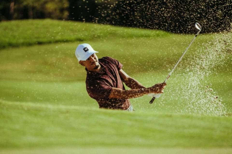Male golfer swings club, sending sand flying on a sunny golf course with lush green grass in the background. Sports and recreation.