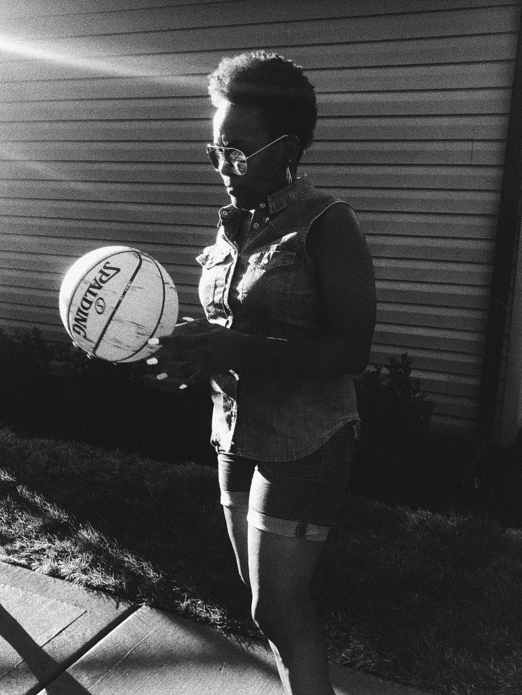 Monochrome medium shot of an adult female in sunglasses and a denim vest outdoors, holding a basketball with a house in the background.