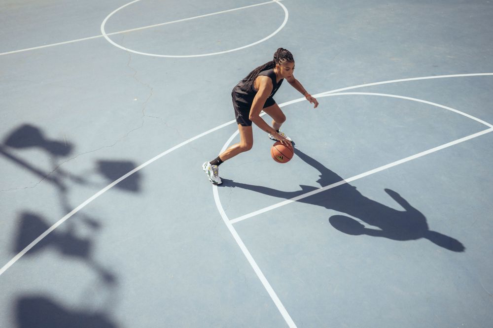 High-angle view of a young woman dribbling a basketball on a blue outdoor court with white lines, casting a long shadow.