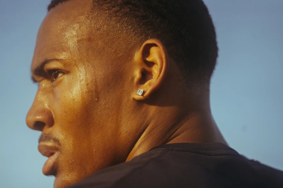 Close-up of a sweaty adult Black male with an earring, illuminated by golden light against a light blue background. Fitness theme.