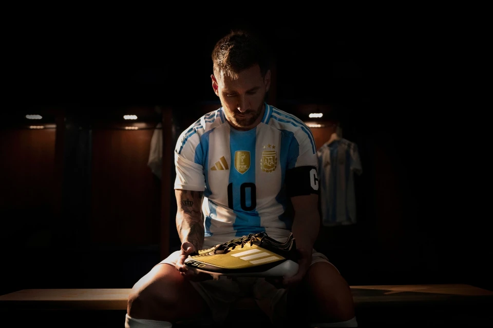 Soccer player Lionel Messi holding a golden Adidas soccer shoe while sitting in a locker room wearing an Argentina jersey.
