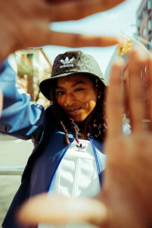 Smiling Black woman in Adidas bucket hat and jacket frames herself with her fingers in an urban environment on a sunny day.