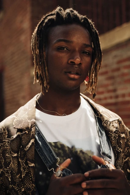 Portrait of a young adult man with dreadlocks wearing a printed T-shirt and jacket with a brick wall background in an outdoor setting.