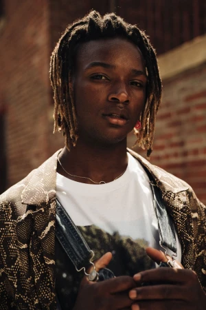 Portrait of a young adult man with dreadlocks wearing a printed T-shirt and jacket with a brick wall background in an outdoor setting.