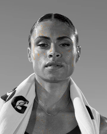 Close-up portrait of a Black female athlete with glistening skin and a towel draped over her shoulders. Features a grayscale background.