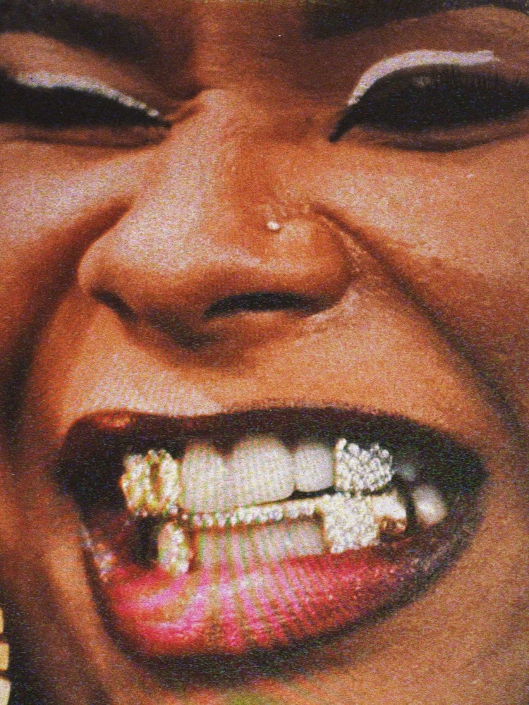 Close-up shot of an African-American woman's face with a smile revealing bejeweled gold and diamond teeth jewelry and a nose piercing.