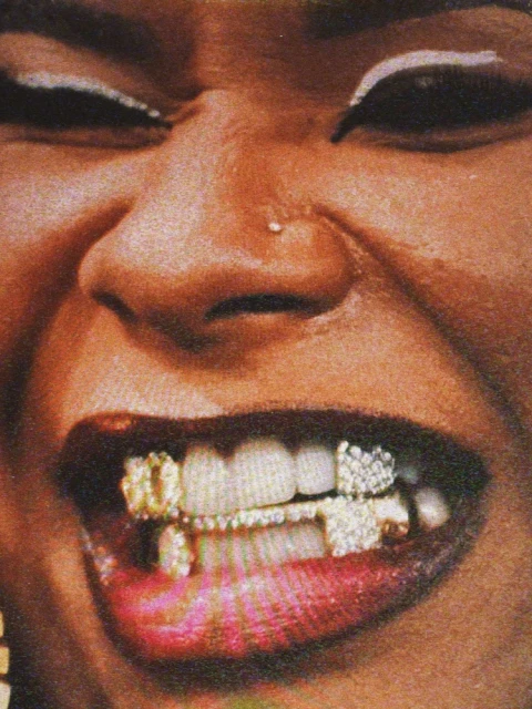 Close-up shot of an African-American woman's face with a smile revealing bejeweled gold and diamond teeth jewelry and a nose piercing.