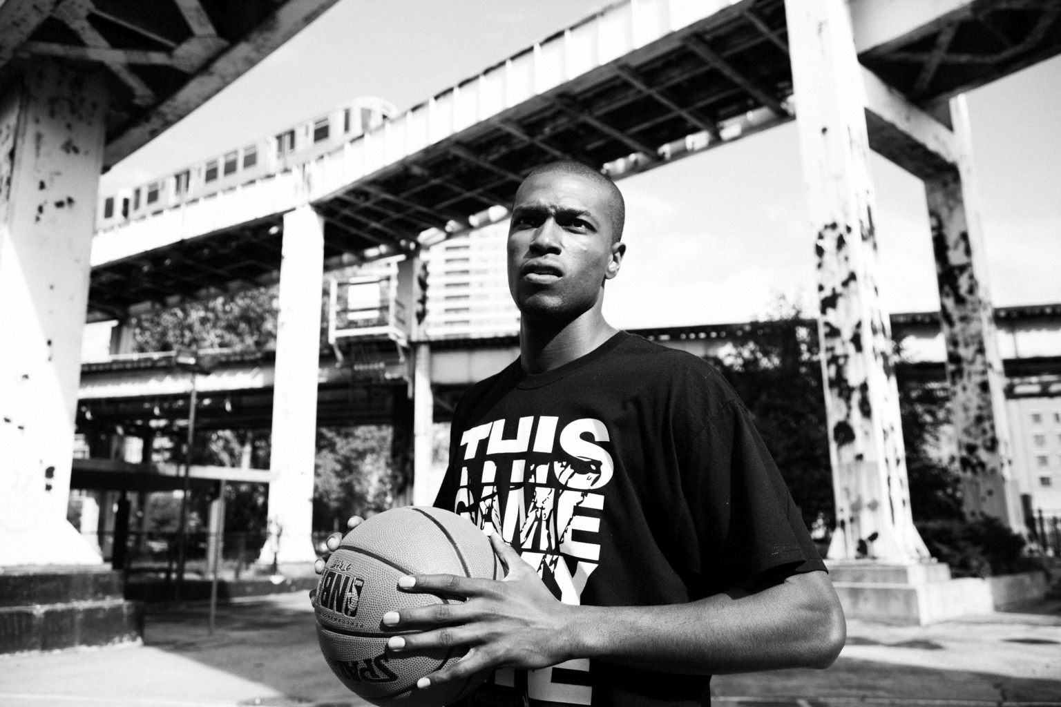 Black and white shot of a man holding a basketball under an overpass with a train passing in the background on a sunny day.