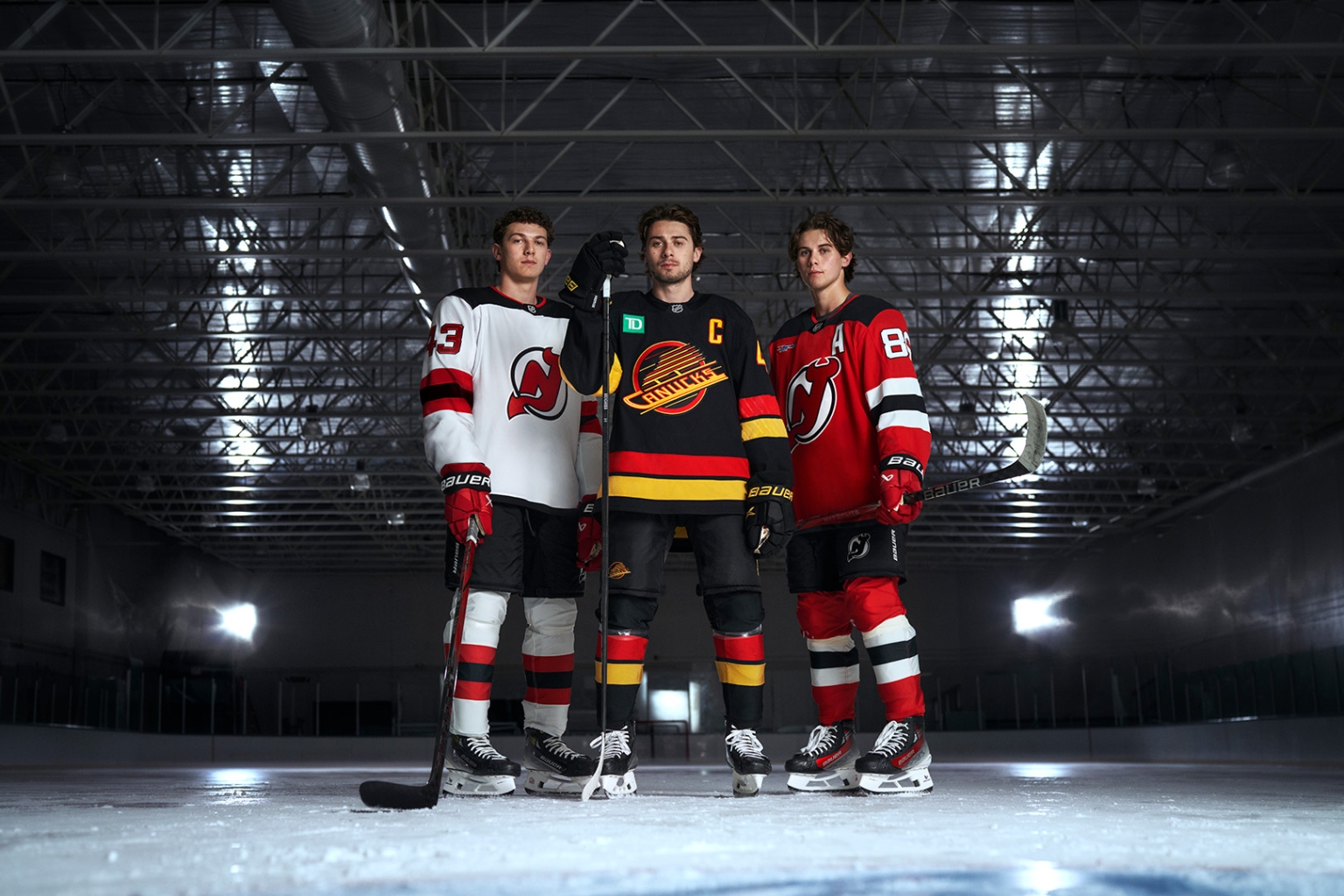 Three male ice hockey players in team jerseys pose on the ice rink, holding hockey sticks, with an indoor stadium visible in the background.