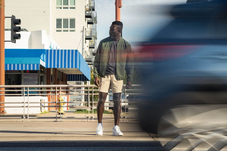 A Black man in khaki shorts and a bomber jacket stands on a city sidewalk next to a street with blurry car in motion.