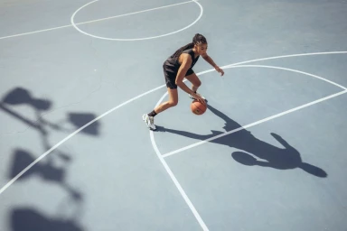 High-angle shot of a female basketball player dribbling a ball on an outdoor court with sharp shadows.