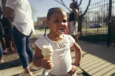 Young child holding an ice cream cone with ice cream on her face and white shirt, photographed outdoors by Marcus Smith