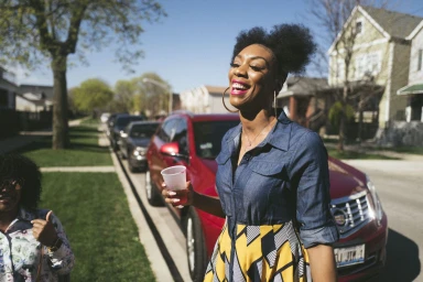 Smiling Black woman in a denim shirt holding a red cup standing on a street in front of a red car