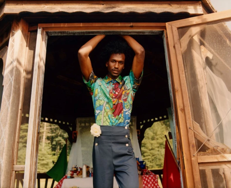 A person stands in a wooden gazebo wearing a green patterned shirt, gray pants, and a flower at the waist against a nature backdrop.