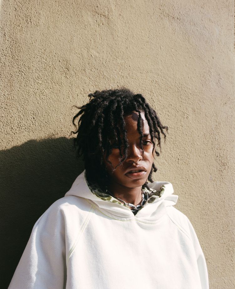 Close-up portrait of an adult with dreadlocks wearing a white hoodie, standing against a tan wall in natural light, outdoor setting.
