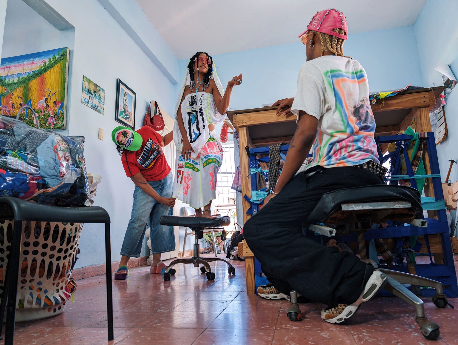 Three people in a room, one sitting in an office chair, one standing on a chair, and one adjusting the dress of the standing woman.