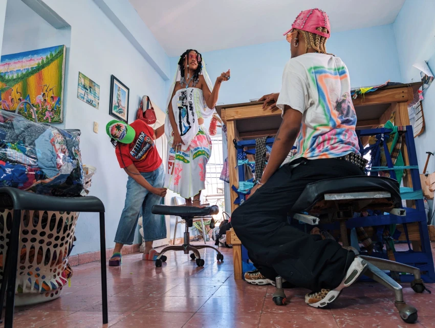Three people in a room, one sitting in an office chair, one standing on a chair, and one adjusting the dress of the standing woman.