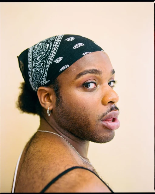 Portrait of a person with dark hair and a bandana, wearing a black tank top and silver jewelry, against a light beige background.
