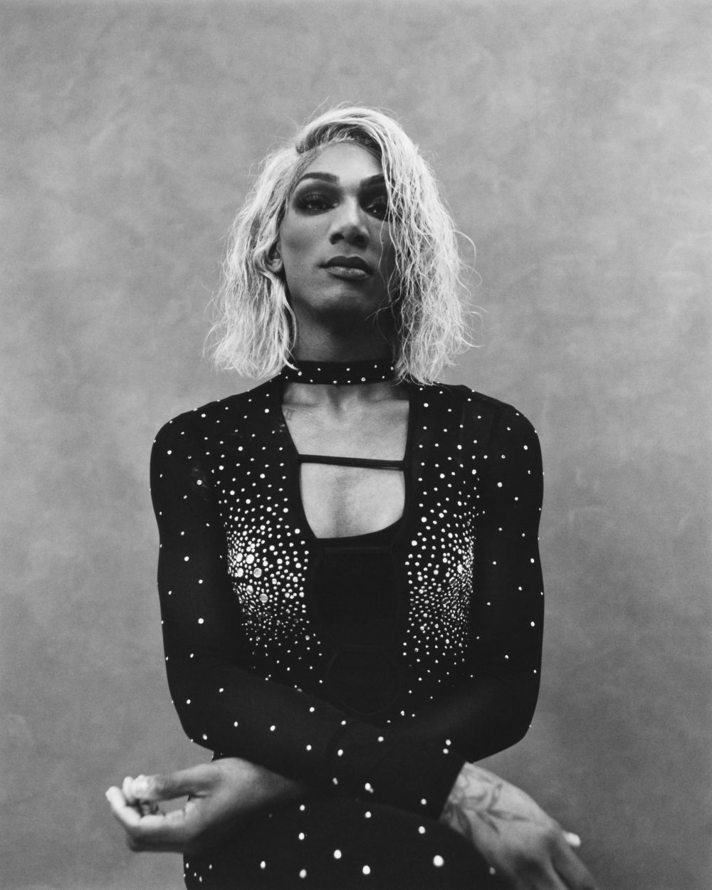 Monochrome portrait of a person with blonde wavy hair, wearing a dark, form-fitting dress with a choker necklace against a textured grey background.