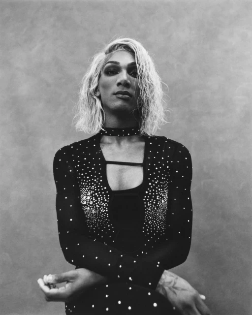 Monochrome portrait of a person with blonde wavy hair, wearing a dark, form-fitting dress with a choker necklace against a textured grey background.