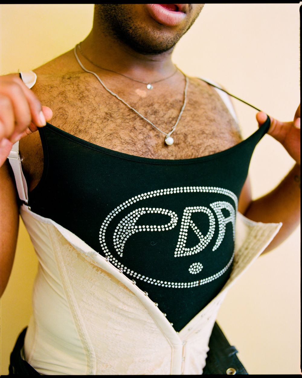 Adult male wearing a black tank top with an emblem and vintage corset around his waist while posing indoors against a neutral background.