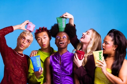 Group of five diverse young adults posing with colorful beauty product containers against a bright blue backdrop in a studio setting.