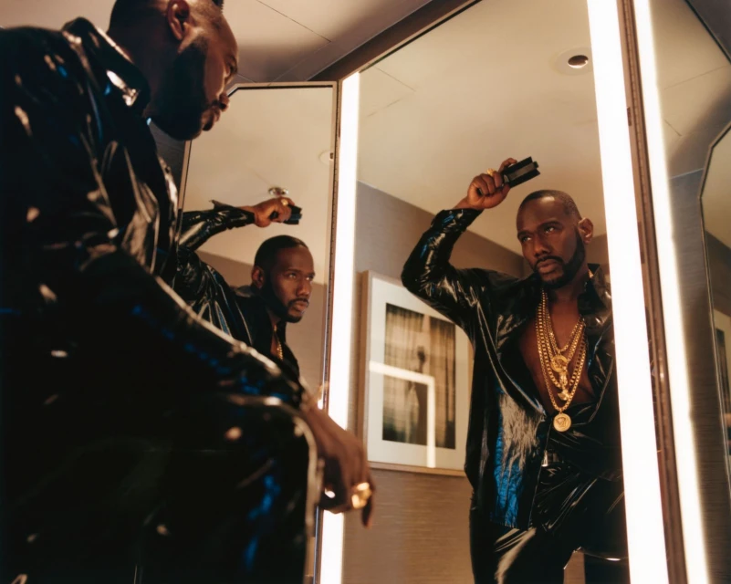 Stylish adult man with gold chains and a shiny black shirt, looking at his reflection in a mirror indoors, holding a comb.