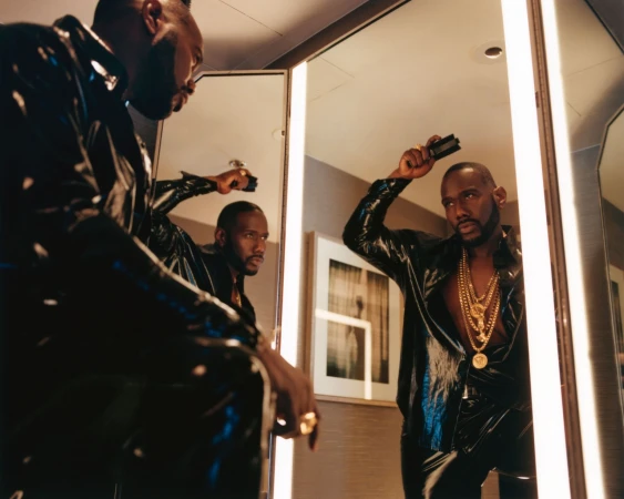 Stylish adult man with gold chains and a shiny black shirt, looking at his reflection in a mirror indoors, holding a comb.