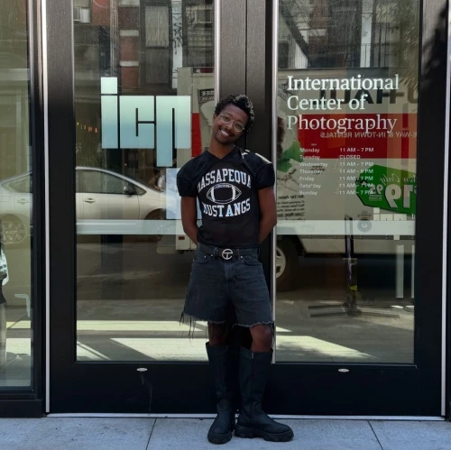 Person poses in front of International Center of Photography wearing Massapequa Mustangs shirt, black shorts and boots on city street.