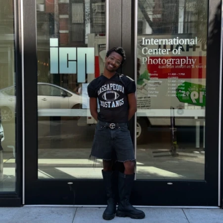 Person poses in front of International Center of Photography wearing Massapequa Mustangs shirt, black shorts and boots on city street.