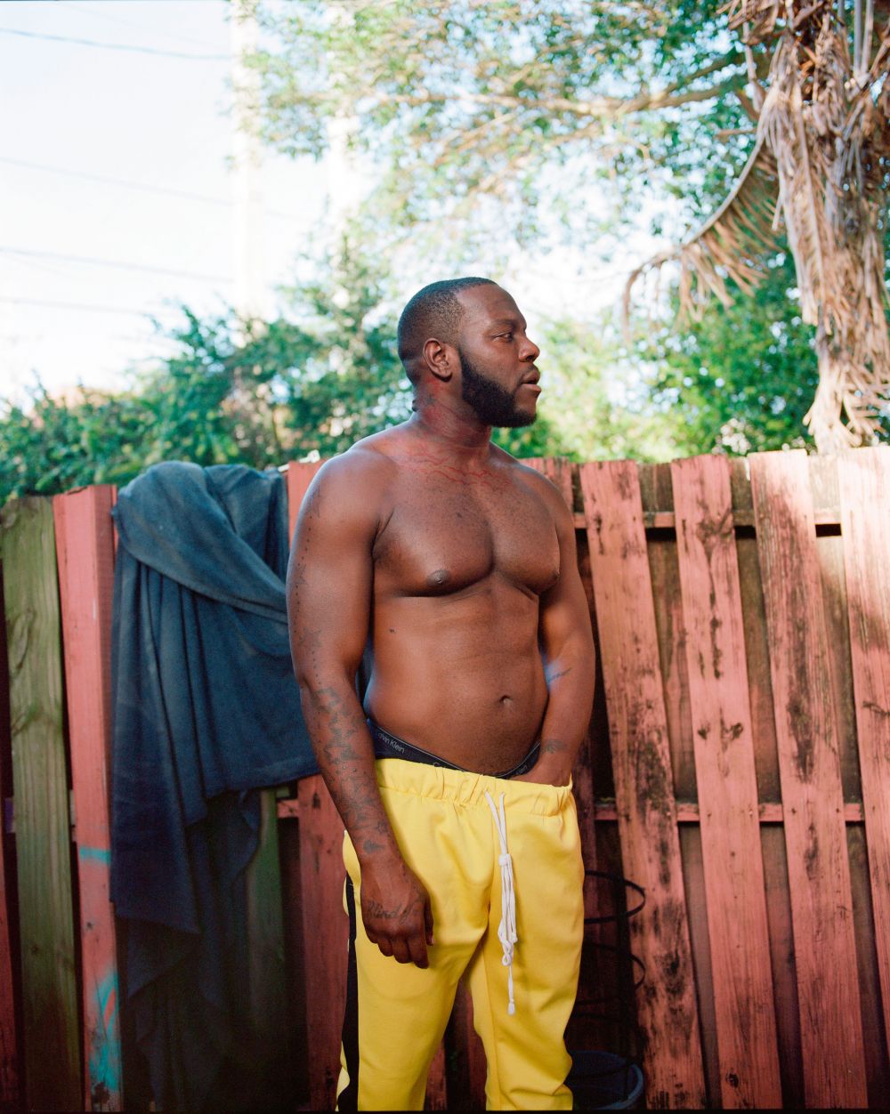 An African-American man with a beard, standing shirtless outdoors with yellow sweatpants, posing in front of a weathered wooden fence.