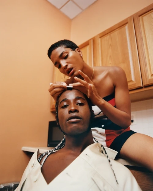 Two adult women in an indoor setting, one seated on a kitchen counter while the other stands behind applying makeup to her forehead.