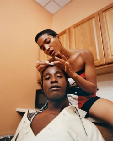 Two adult women in an indoor setting, one seated on a kitchen counter while the other stands behind applying makeup to her forehead.