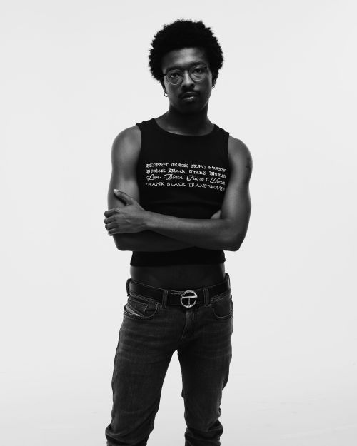 Portrait of a Black man with glasses and short afro wearing a black tank top and denim jeans with arms crossed against a white background.