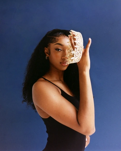 Portrait of a Black woman with curly hair and a black top holding a gold decorative object in front of her face against a blue background.