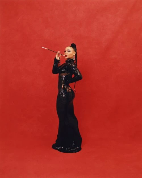 Adult woman in a floor-length sequined gown, posing in a photo studio with a red backdrop, and holding a cigarette holder.