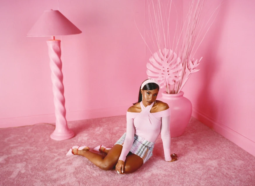 Young woman poses on a pink carpet in front of a pink vase and palm leaves, matching pink walls, and a pink floor lamp in a studio.