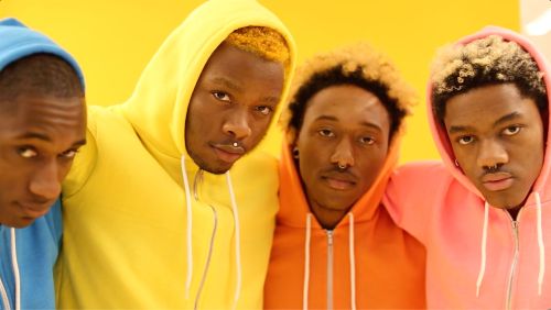 A group of four Black men in colorful hoodies pose close together against a bright yellow background in a studio setting. The photo is a medium shot.