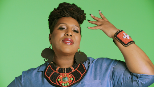 Portrait of an adult woman with dreadlocks and beaded jewelry in a photo studio, striking a pose against a green backdrop.