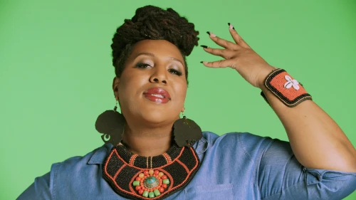 Portrait of an adult woman with dreadlocks and beaded jewelry in a photo studio, striking a pose against a green backdrop.