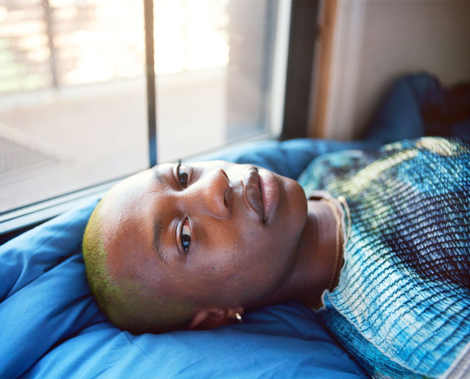 Close-up of a black adult female with dyed green hair, lying on blue bedding, looking up toward the camera by a window.