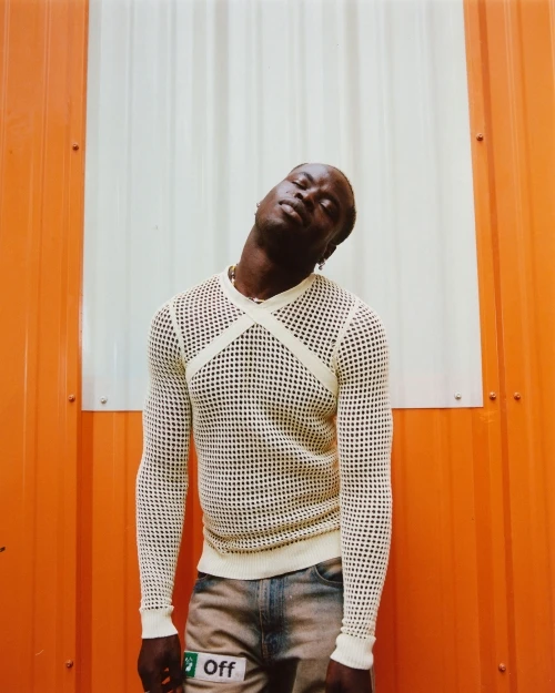 A young adult African male stands in front of an orange and white wall, wearing a white mesh shirt and jeans. The man is stylish.