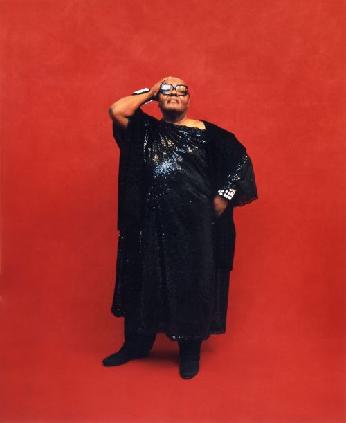 Full-length portrait of a Black adult wearing a sequined dress and glasses, hand on head, posing against a vibrant red background.
