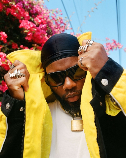 Black man wearing black durag, sunglasses, white t-shirt, and yellow jacket with multiple silver rings, photographed by Myles Loftin