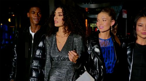 Group of four multi-ethnic young adults in fashionable leather jackets and dresses walk together outdoors at night, with dark urban background.