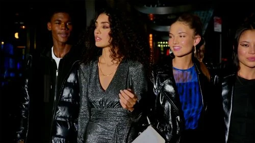 Group of four multi-ethnic young adults in fashionable leather jackets and dresses walk together outdoors at night, with dark urban background.